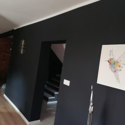 Dar-but firma handlowo usługowa Dariusz Czajkowski - Pomieszczenie z czarnymi ścianami, białym sufitem i listwami przypodłogowymi, dekorowane zegarem, obrazem z kolibrem i metalową rzeźbą, widoczne schody w tle.