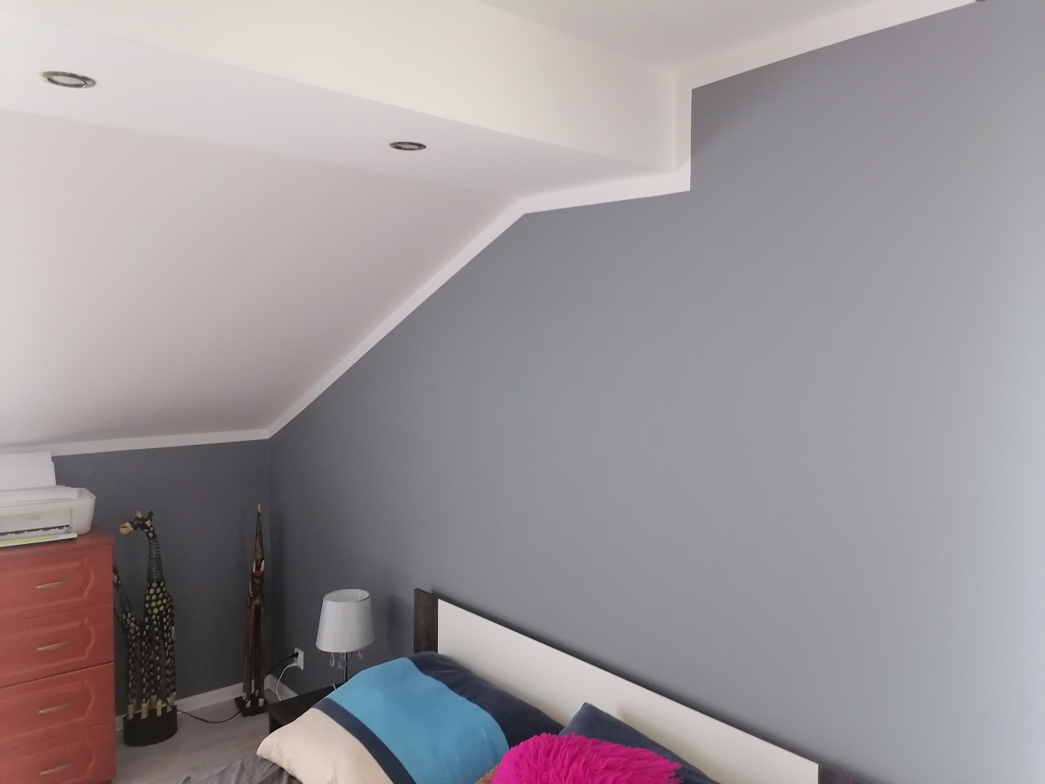 Pokój na poddaszu po odświeżeniu ścian szarą farbą, widoczne skosy sufitu, oświetlenie punktowe i fragment sypialni z łóżkiem oraz dekoracyjnymi figurkami.