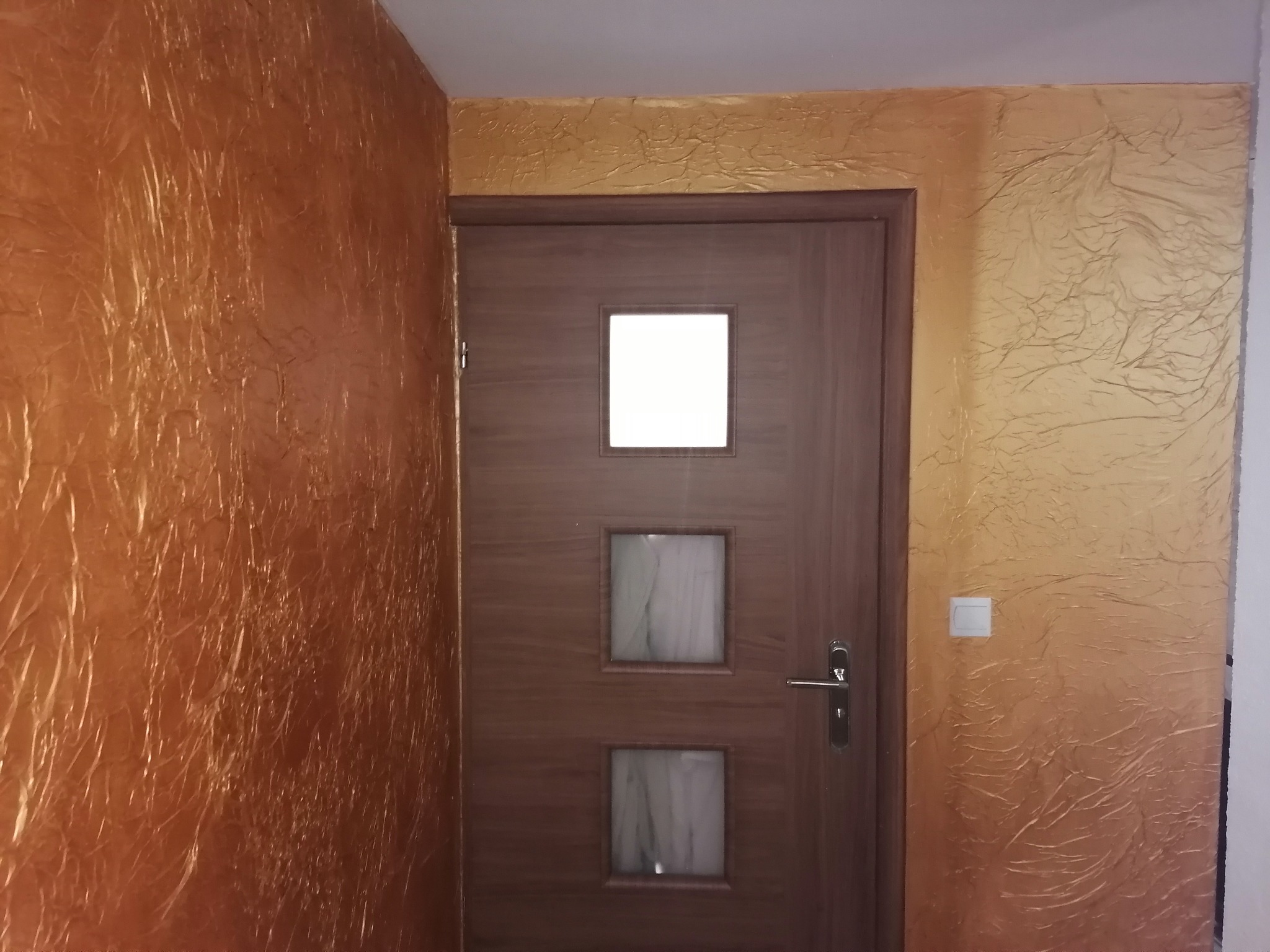 Brązowe drzwi z trzema kwadratowymi okienkami na tle ściany pokrytej dekoracyjną, złoto-brązową fakturą przypominającą pomarszczony papier.
