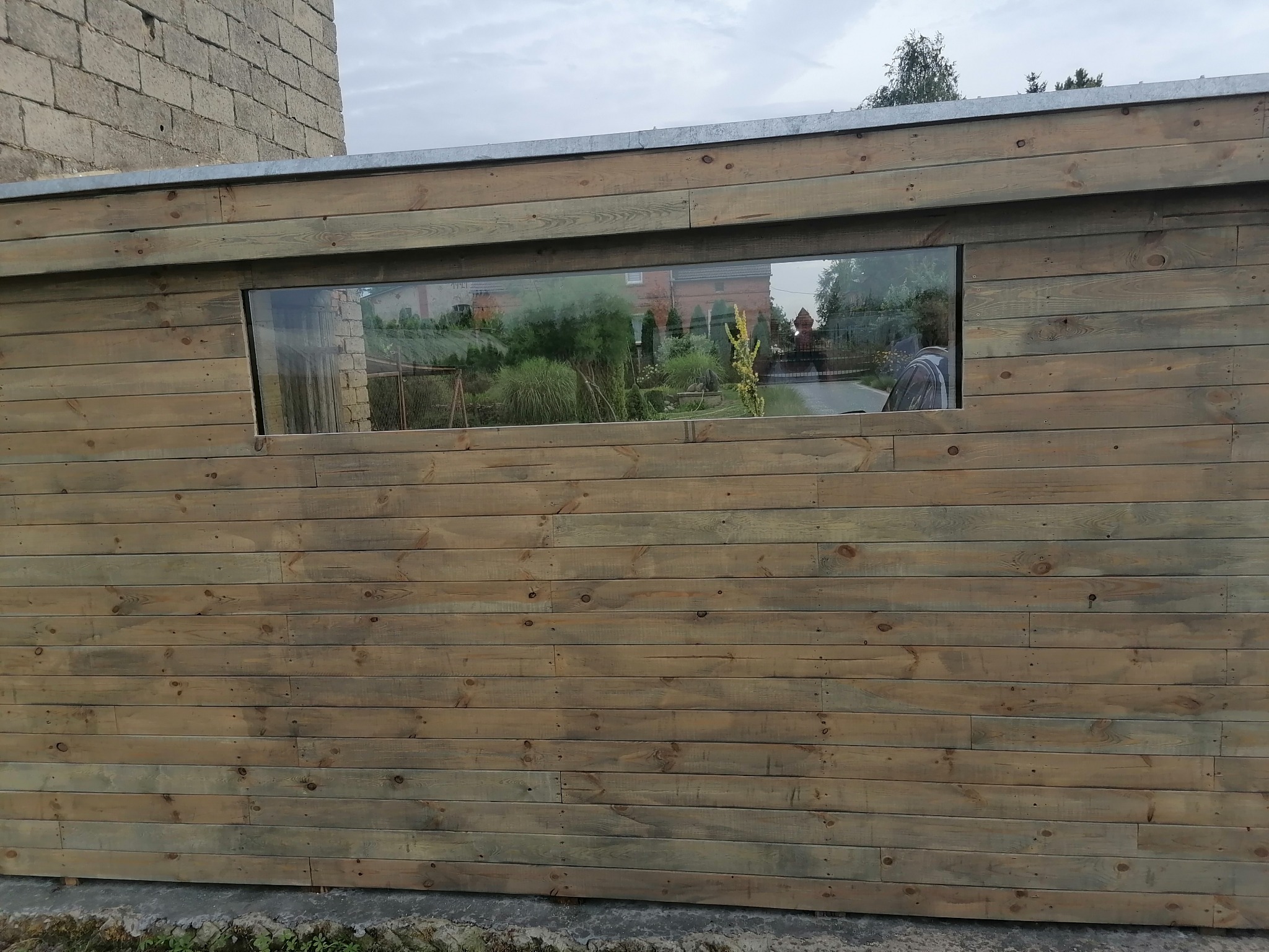 Drewniana ściana z poziomych desek, z prostokątnym oknem odbijającym zieleń ogrodu i budynek w tle.