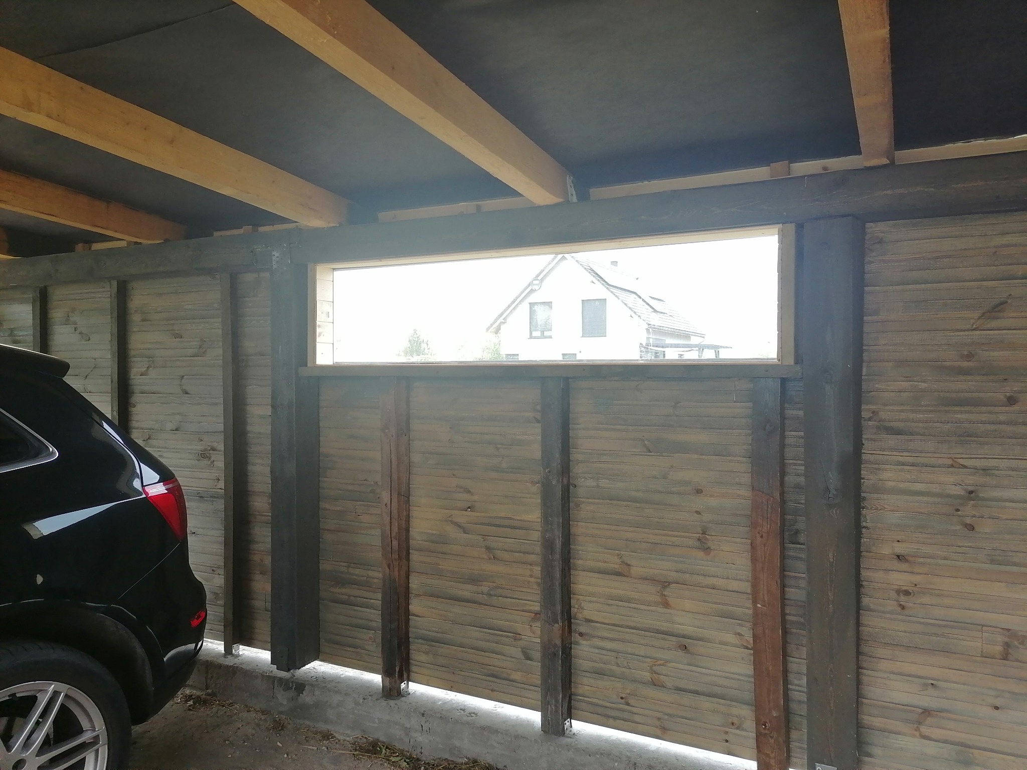 Fragment konstrukcji z drewnianych paneli w Górze, z widocznym oknem i częścią samochodu. Konstrukcja z belek i desek, widok na dom przez okno.
