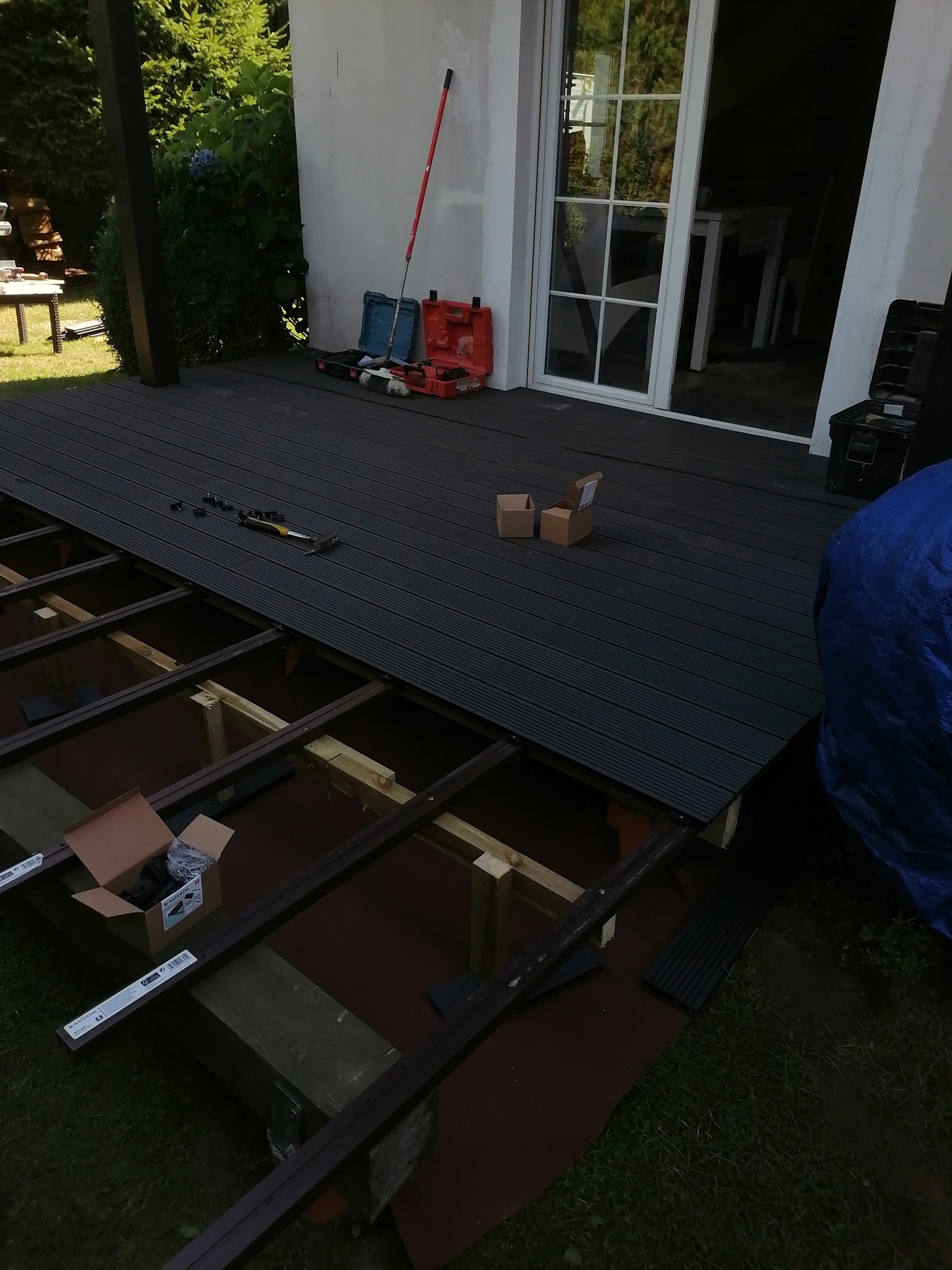 Montaż ciemnej deski kompozytowej na tarasie, widoczne legary, narzędzia i akcesoria montażowe, częściowo ukończona powierzchnia, skrzynki narzędziowe w tle.