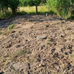 Teren po przygotowaniu pod instalację, widoczne dwa pionowe przewody wentylacyjne i okrągła pokrywa włazu, otoczone świeżo przekopaną ziemią, w tle zieleń drzew i krzewów.