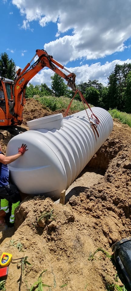 Pomarańczowa koparka JCB opuszcza biały, żebrowany zbiornik do wykopu w ziemi, widoczny fragment pracownika w odblaskowej kamizelce i czerwonej rękawicy.