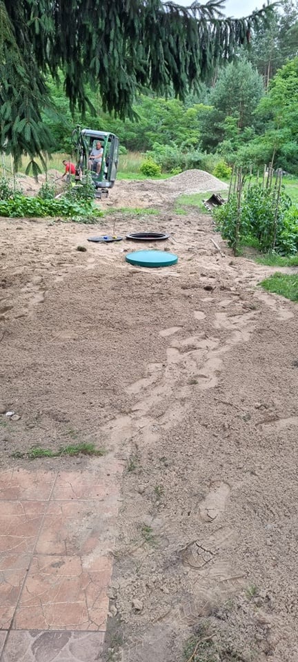 Teren po wykonanych pracach ziemnych, widoczne włazy do zbiorników, mini koparka z operatorem w oddali, świeżo nawieziona ziemia, ślady butów na ziemi.