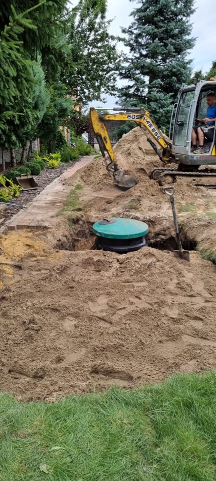 Żółta mini koparka zasypuje piaskiem zielony zbiornik w wykopie, obok stoi łopata, w tle widoczny zadbany ogród z kostką brukową.