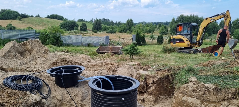 Montaż przydomowej oczyszczalni ścieków w Radomiu: widok na dwa czarne zbiorniki połączone rurą, wykop, żółta koparka i wózek budowlany na tle zielonego krajobrazu.