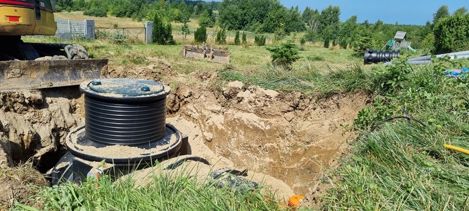 Częściowo zakopany zbiornik przydomowej oczyszczalni ścieków w wykopie, widoczny fragment koparki i elementy instalacji na tle zieleni i drzew.