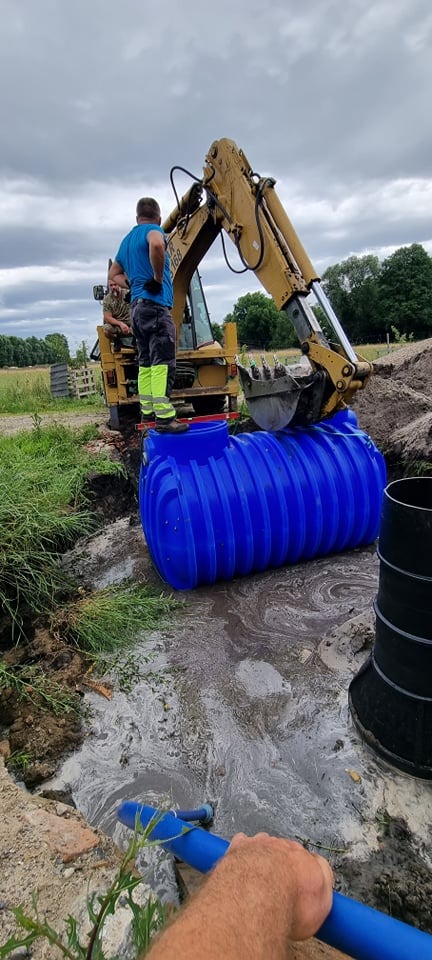 Żółta koparka umieszcza niebieski zbiornik z tworzywa sztucznego w wykopie, dwóch mężczyzn nadzoruje proces, w tle zielona trawa i pochmurne niebo.
