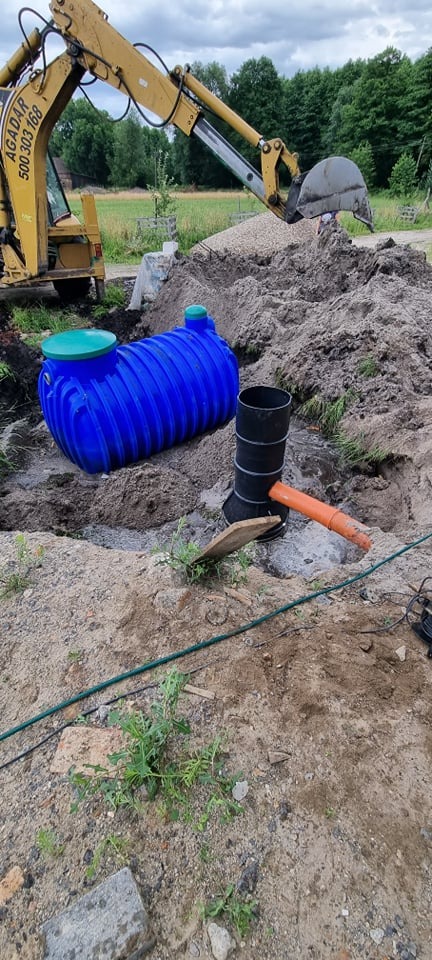 Niebieski zbiornik przydomowej oczyszczalni ścieków leży w wykopie, obok czarnej rury kanalizacyjnej i pomarańczowego przewodu, z koparką w tle, podczas prac ziemnych.
