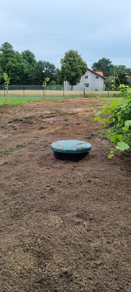 Zielona pokrywa włazu przydomowej oczyszczalni ścieków wystająca z ziemi na świeżo wyrównanym terenie, w tle dom jednorodzinny i drzewa.