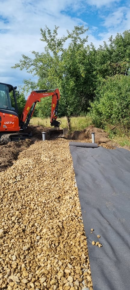 Pomarańczowy minikoparka Kubota U27-4 przy wykopie z dwoma pionowymi rurami, podłoże częściowo wyłożone żwirem i czarną geowłókniną, w tle zieleń drzew i krzewów.