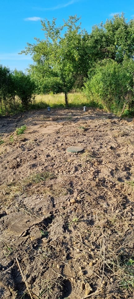 Teren po przygotowaniu pod instalację, widoczne dwa pionowe przewody wentylacyjne i okrągła pokrywa włazu, otoczone świeżo przekopaną ziemią, w tle zieleń drzew i krzewów.