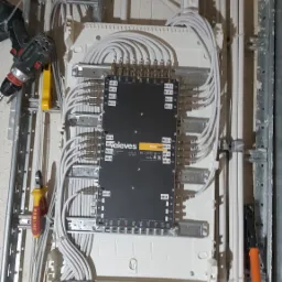 Rozdzielnica elektryczna Televes z licznymi przewodami podłączonymi do złączek, widoczne narzędzia: wiertarka i kombinerki, montaż na szynach metalowych na tle surowej ściany.