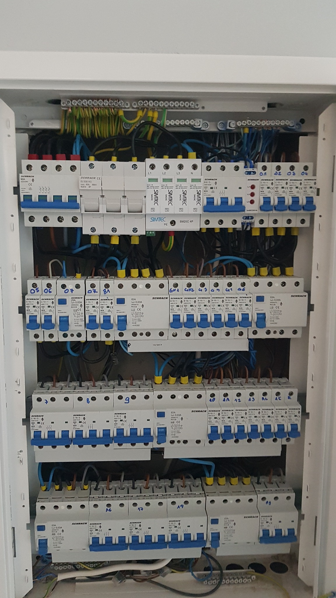 Rozdzielnica elektryczna z bezpiecznikami Schrack i Simtec, widoczne okablowanie i oznaczenia obwodów, wnętrze szafki w kolorze białym.