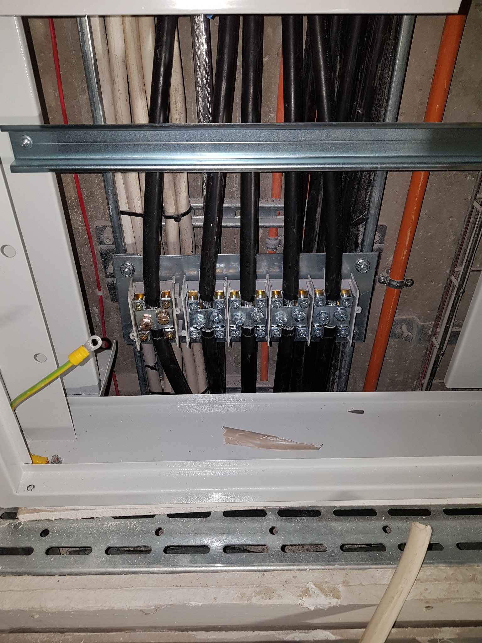 Szczegółowe ujęcie skrzynki elektrycznej z widocznymi przewodami podłączonymi do listwy zaciskowej, przewód uziemiający z żółto-zieloną izolacją po lewej stronie, widoczne rury instalacyjne w tle.