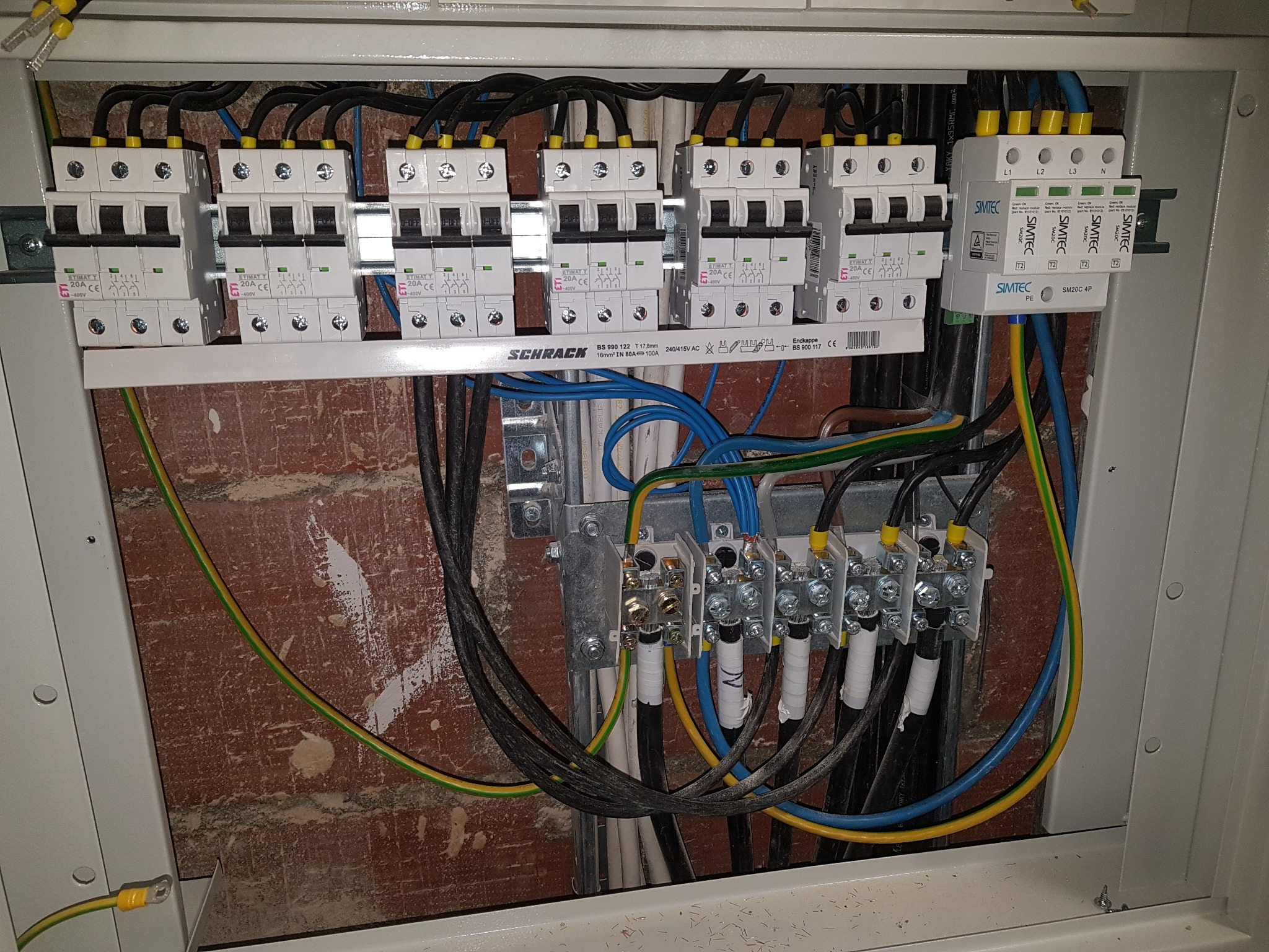 Wnętrze skrzynki elektrycznej z zainstalowanymi bezpiecznikami ETIMATY, ogranicznikiem przepięć SIMTEC i podłączonymi przewodami o różnych kolorach, zamontowanej na ceglanej ścianie.