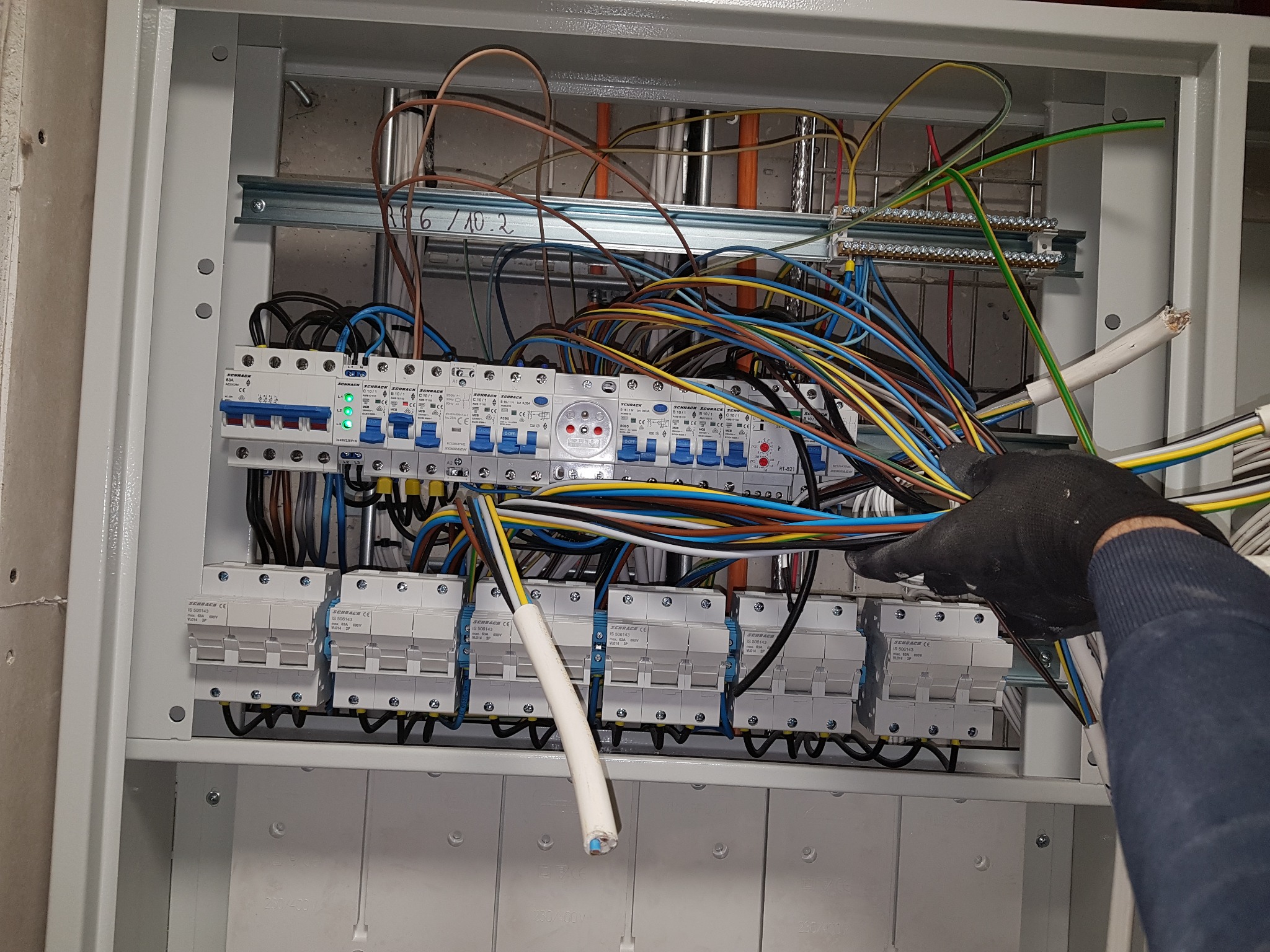 Otwarta skrzynka elektryczna z zamontowanymi bezpiecznikami, zabezpieczeniami przepięciowymi i licznymi przewodami podłączanymi przez elektryka w rękawicy ochronnej.