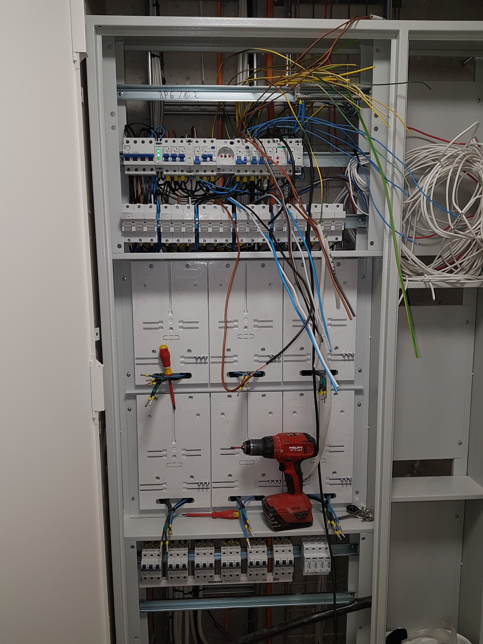 Otwarta szafa elektryczna w trakcie montażu, widoczne okablowanie, bezpieczniki, wkrętarka Hilti i śrubokręty, przewody w różnych kolorach.