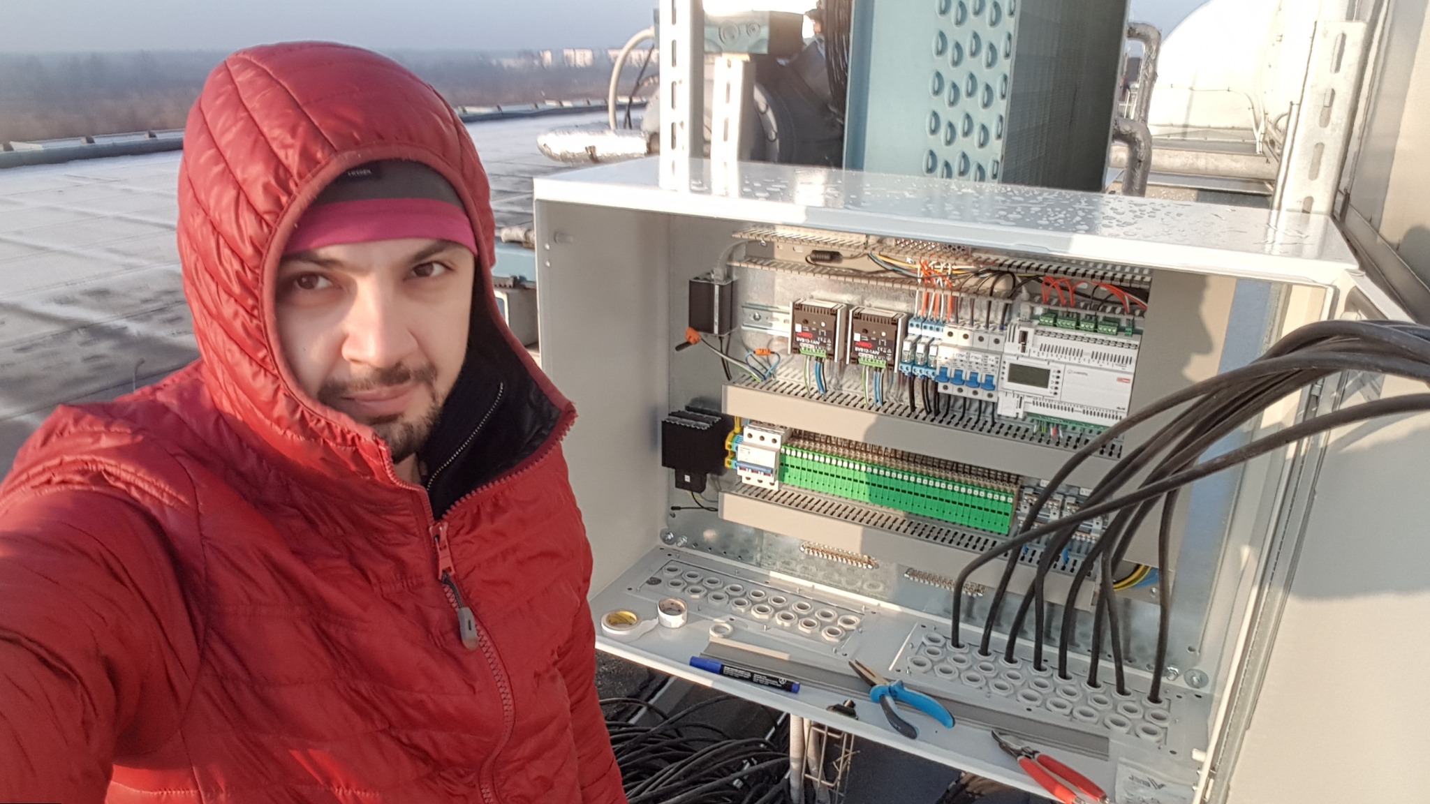 Elektryk w czerwonej kurtce prezentuje otwartą szafę sterowniczą na dachu, z widocznymi przewodami, listwami zaciskowymi i narzędziami pracy.