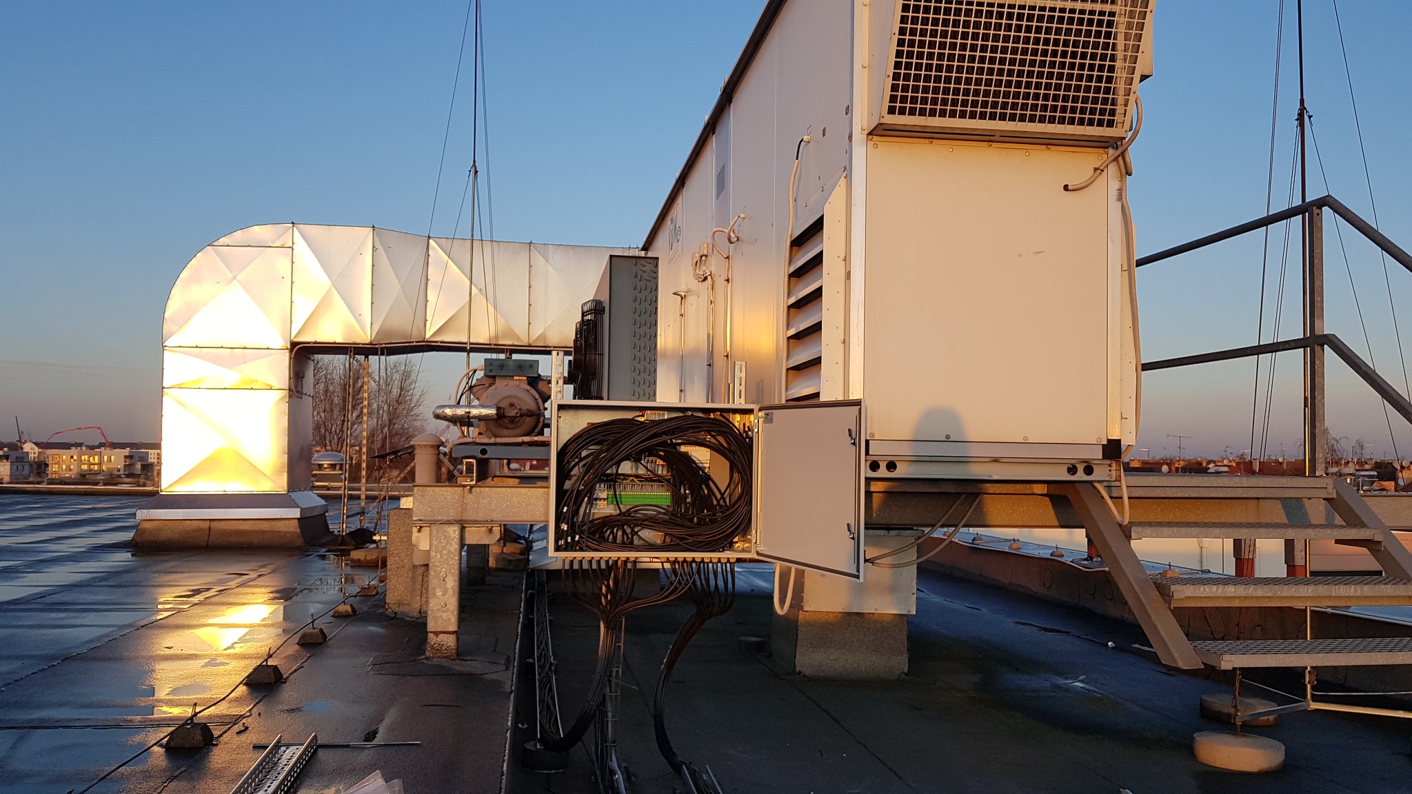 Instalacja przemysłowa na dachu budynku z otwartą skrzynką elektryczną i plątaniną kabli, oświetlona ciepłym światłem zachodzącego słońca, widoczny komin wentylacyjny i drabina dostępu.