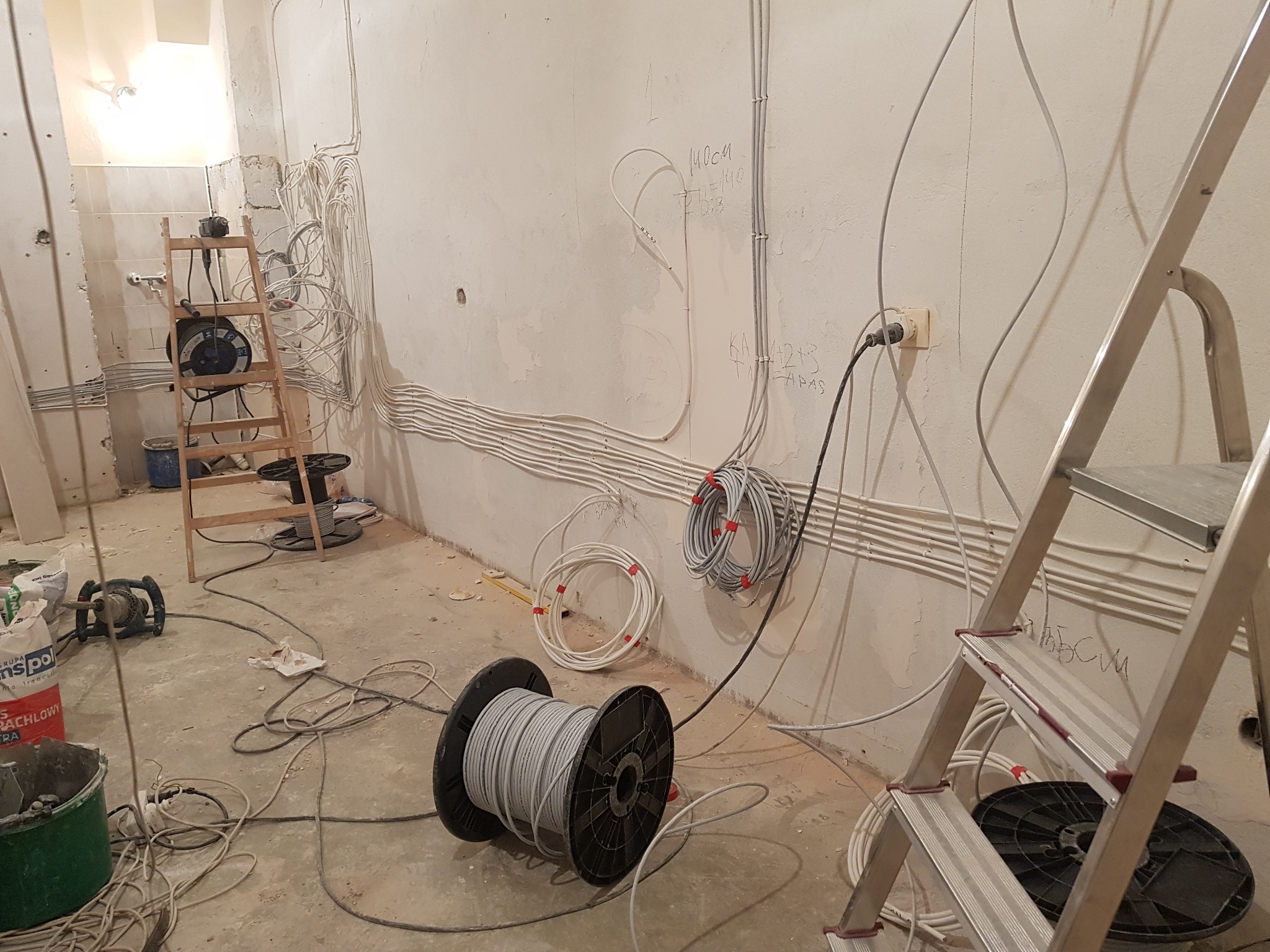 Wnętrze w trakcie prac elektrycznych: widoczne kable prowadzone po ścianach, puszki elektryczne, drabina aluminiowa, drewniana drabina, szpule z kablami na podłodze oraz narzędzia i materiały...