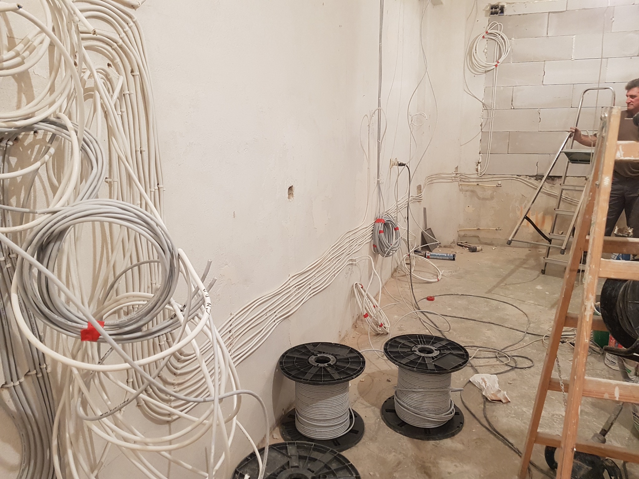 Wnętrze w trakcie remontu z widocznymi wiązkami przewodów elektrycznych przytwierdzonymi do ściany, szpulami z kablami na podłodze i mężczyzną na drabinie w tle.