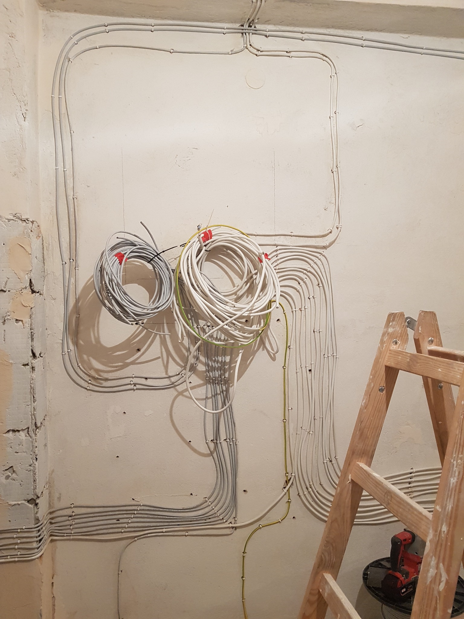 Ściana w trakcie remontu z widocznymi, nowo poprowadzonymi przewodami elektrycznymi w białych peszlach, zwinięte kable na ścianie i drabina z wiertarką w tle.