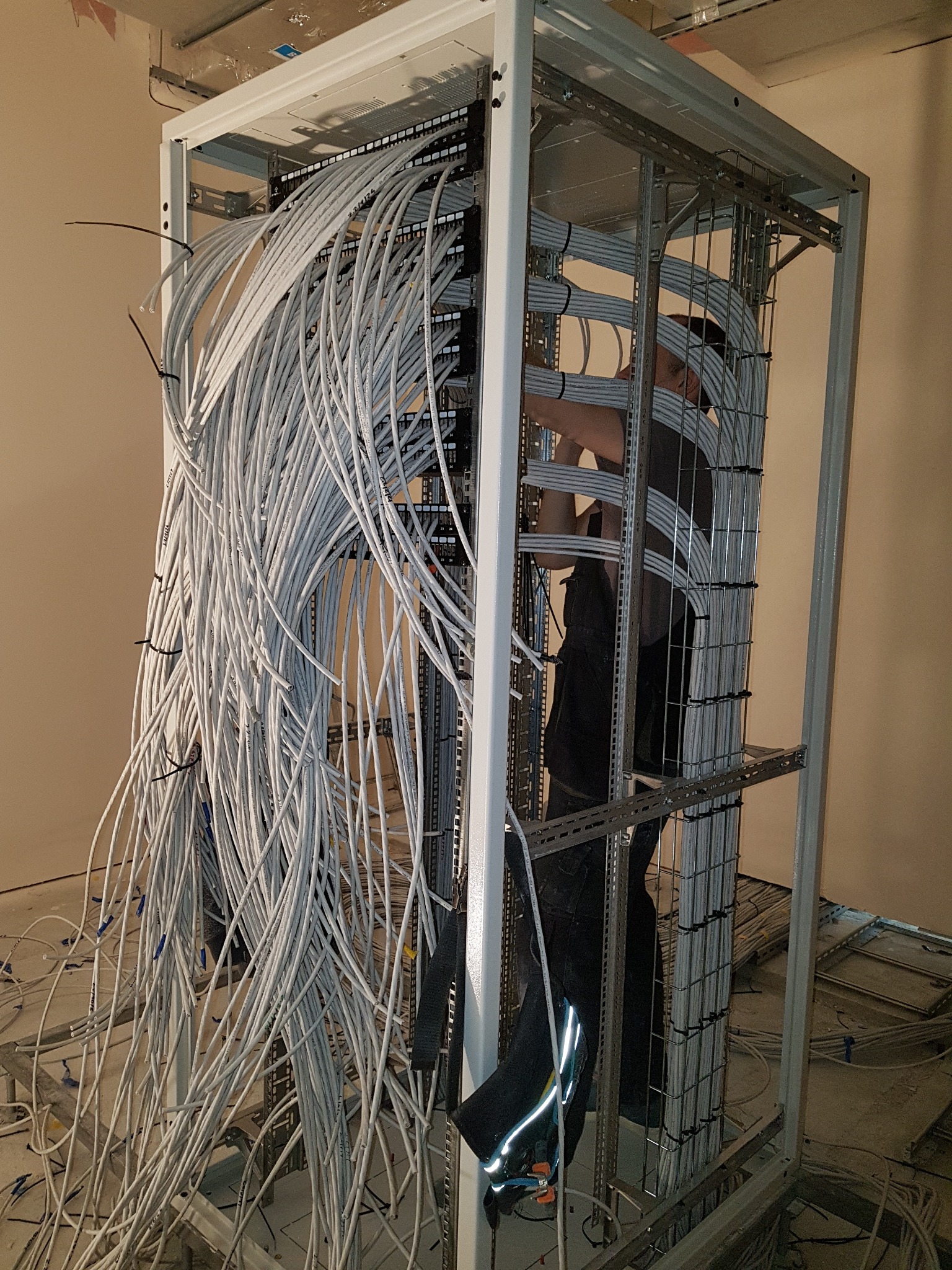 Instalacja okablowania strukturalnego w metalowej szafie rack: widoczne liczne kable kategorii 5e/6 układane przez technika, złączone w panel krosowniczy w pomieszczeniu w stanie surowym.
