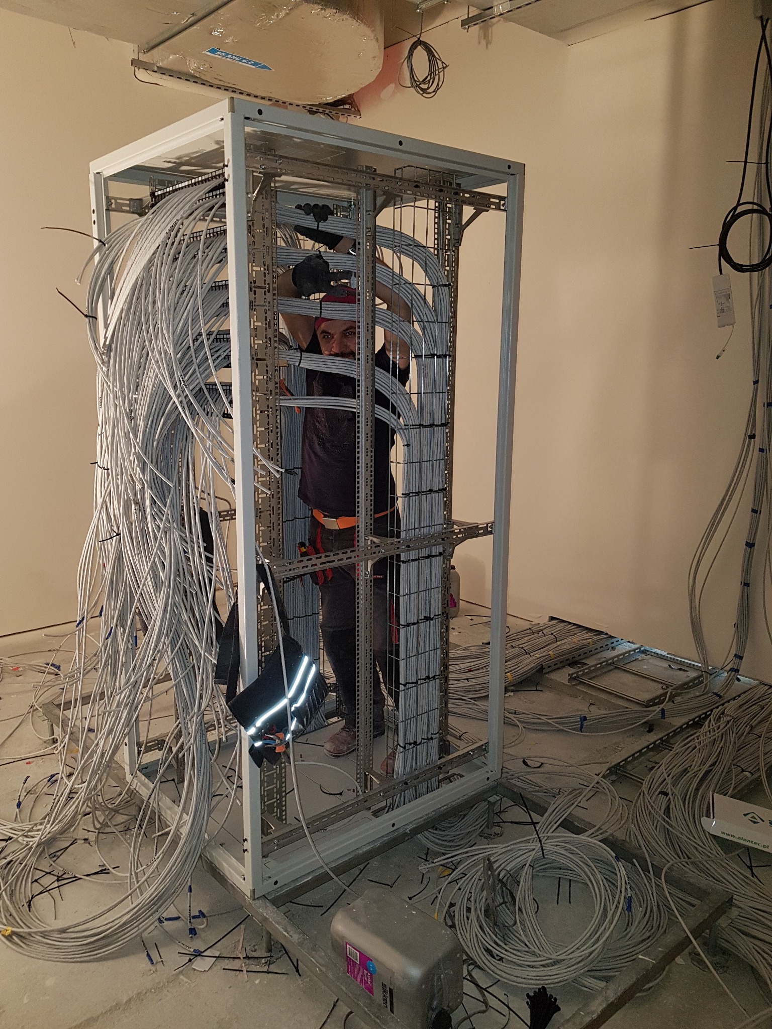 Elektryk w kasku i rękawicach układa kable w metalowej szafie rack, widoczne liczne przewody i elementy instalacji elektrycznej w surowym stanie.