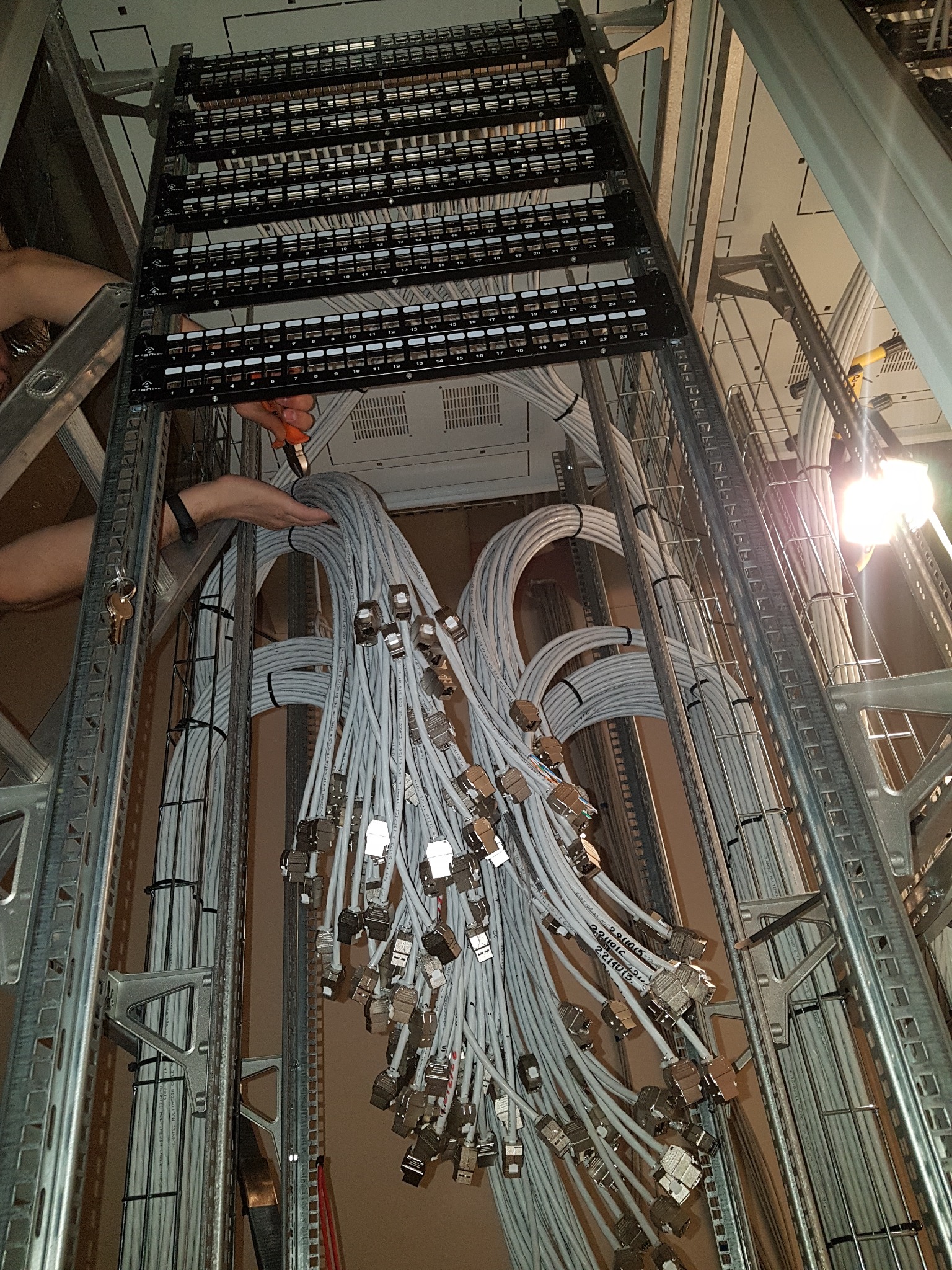 Instalacja patch panela z licznymi kablami zakończonymi wtykami RJ45, widok z dołu, w tle metalowa konstrukcja stelaża, ręce instalatora przy użyciu obcęgów.