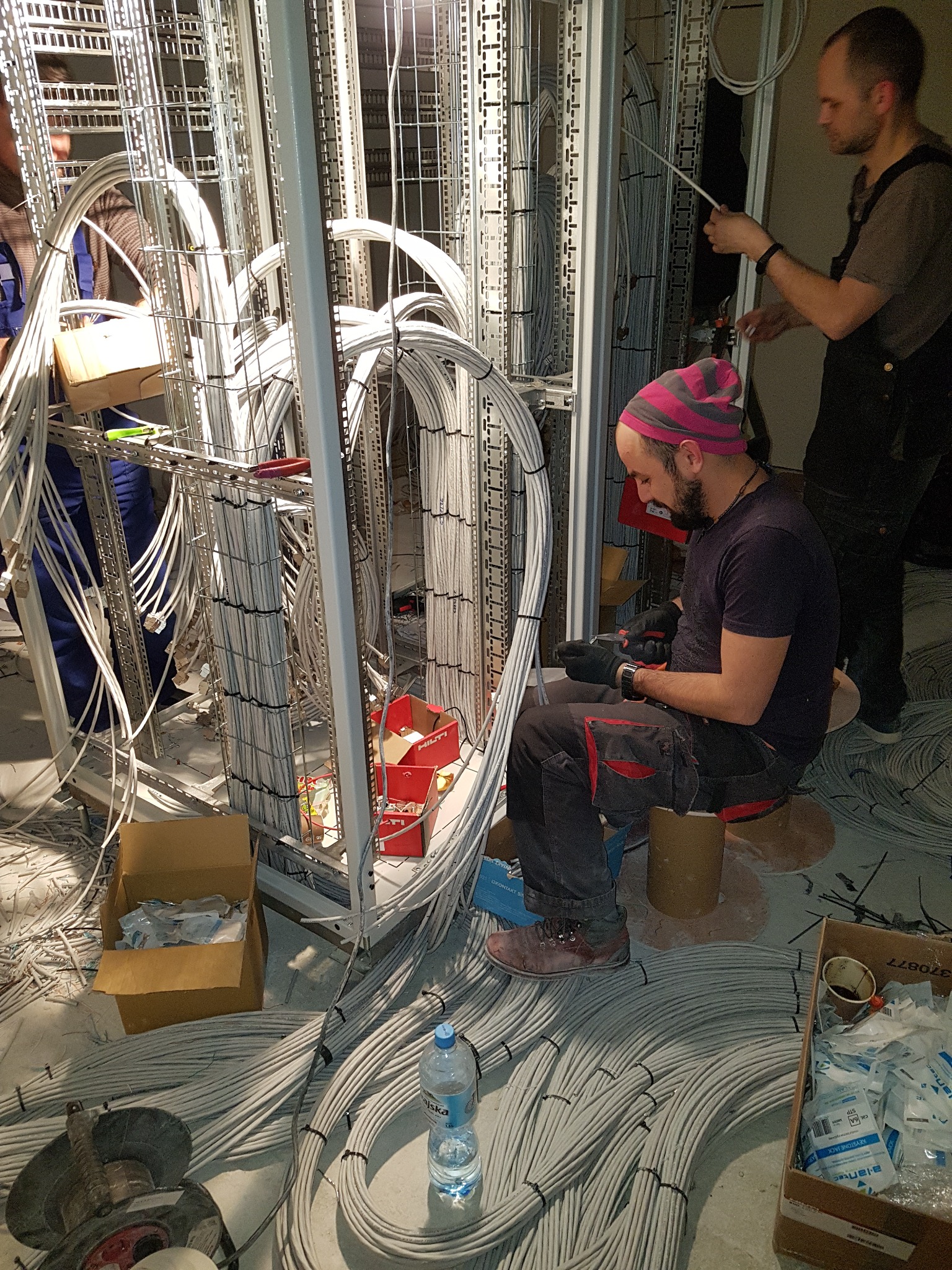 Instalator w czapce w różowe paski siedzi na szpuli kabla, podłączając kable do metalowej ramy w pomieszczeniu z dużą ilością kabli na podłodze. Widoczne narzędzia, pudełka i butelka wody.