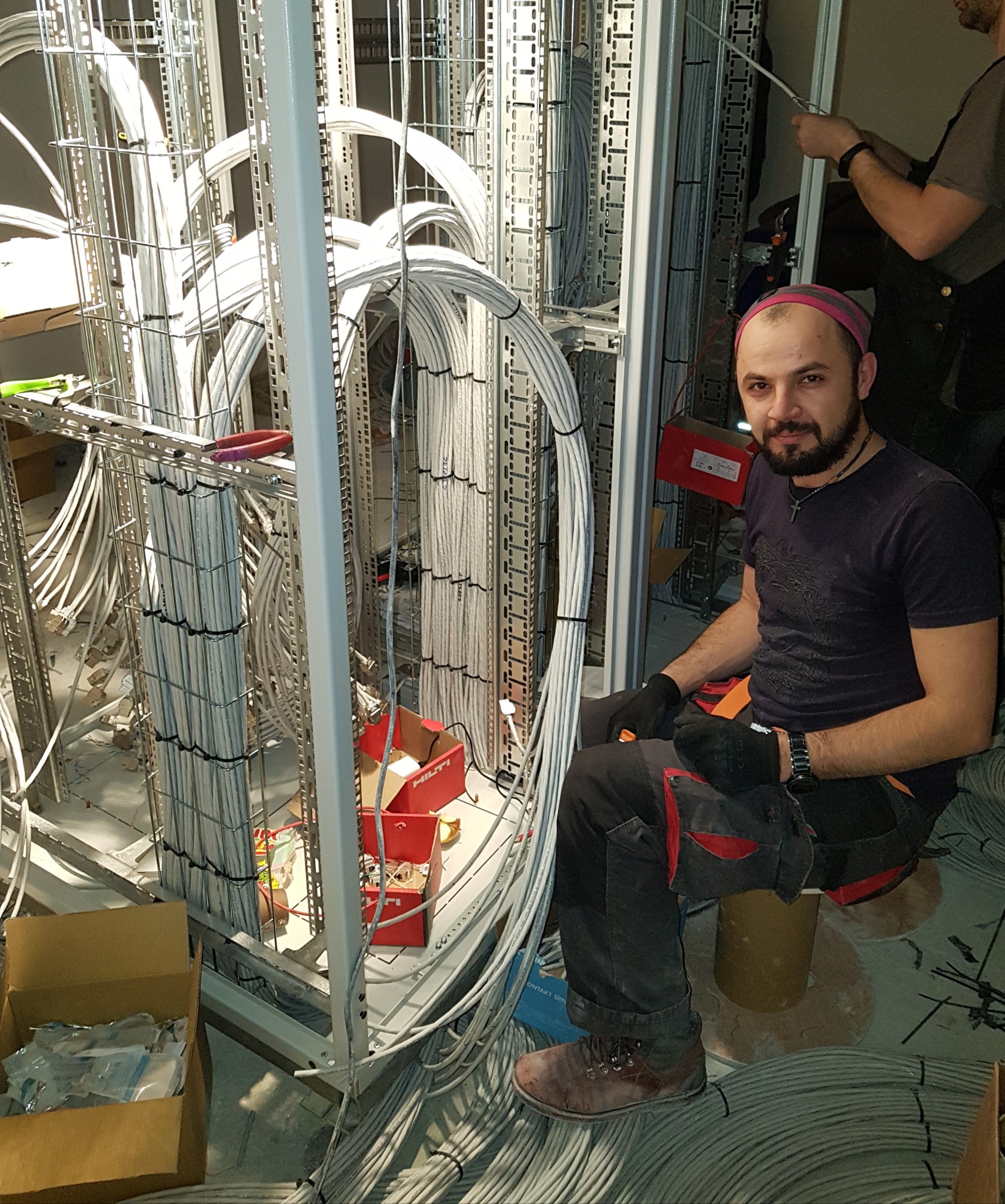 Elektryk z Piły siedzi na bębnie kablowym pośród uporządkowanych wiązek przewodów w metalowej konstrukcji, podczas gdy drugi elektryk pracuje w tle.