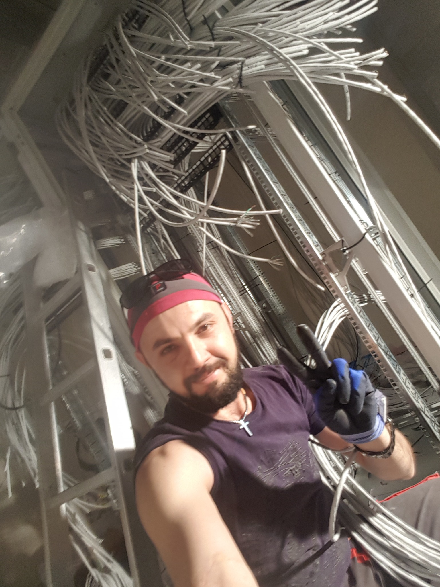 Instalator robi selfie na drabinie, pokazując gest zwycięstwa, z widoczną plątaniną białych kabli pod sufitem na metalowej konstrukcji.