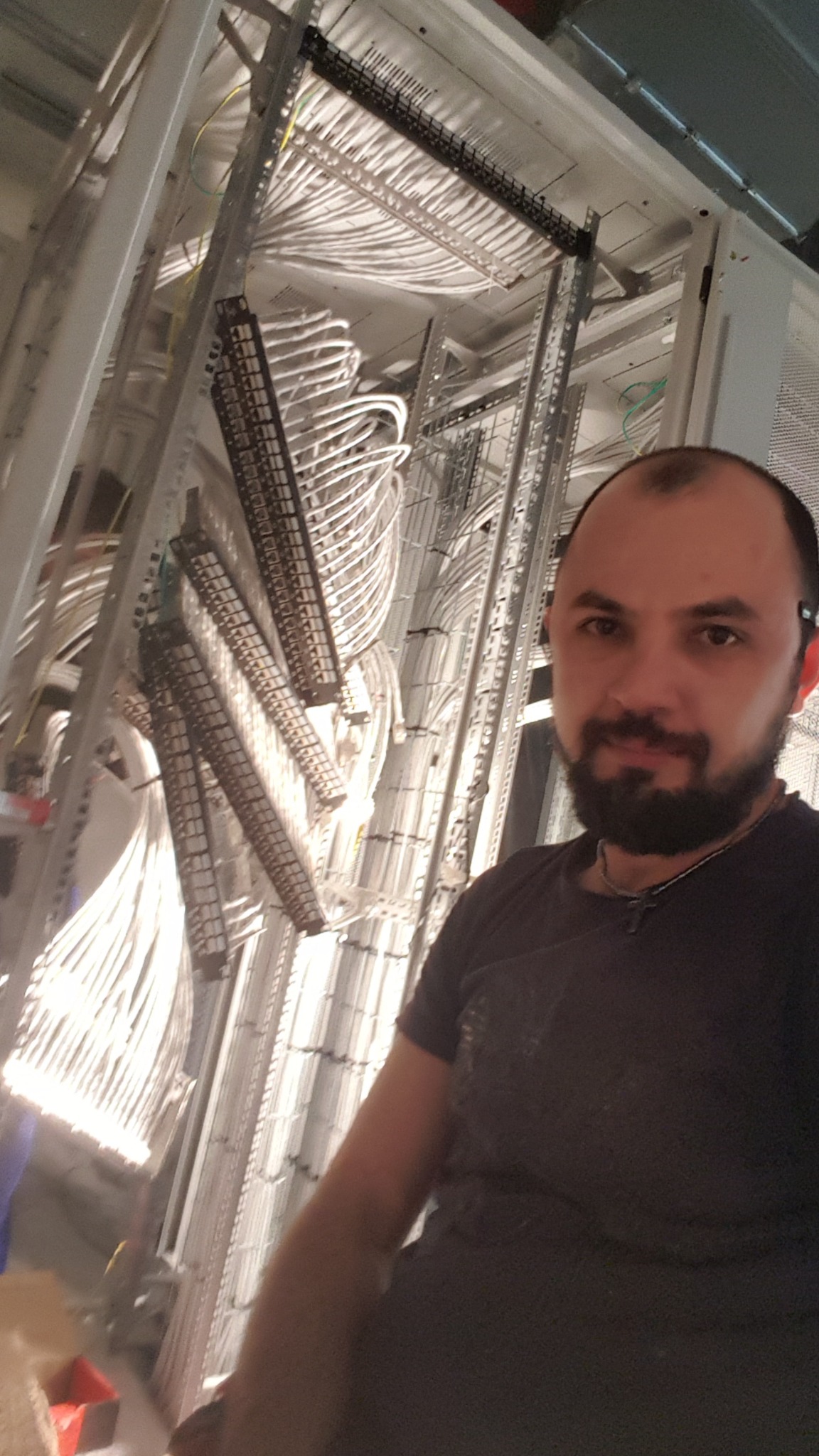 Uśmiechnięty mężczyzna na tle rozbudowanej instalacji kablowej w szafie serwerowej, widoczne liczne kable uporządkowane w korytkach.