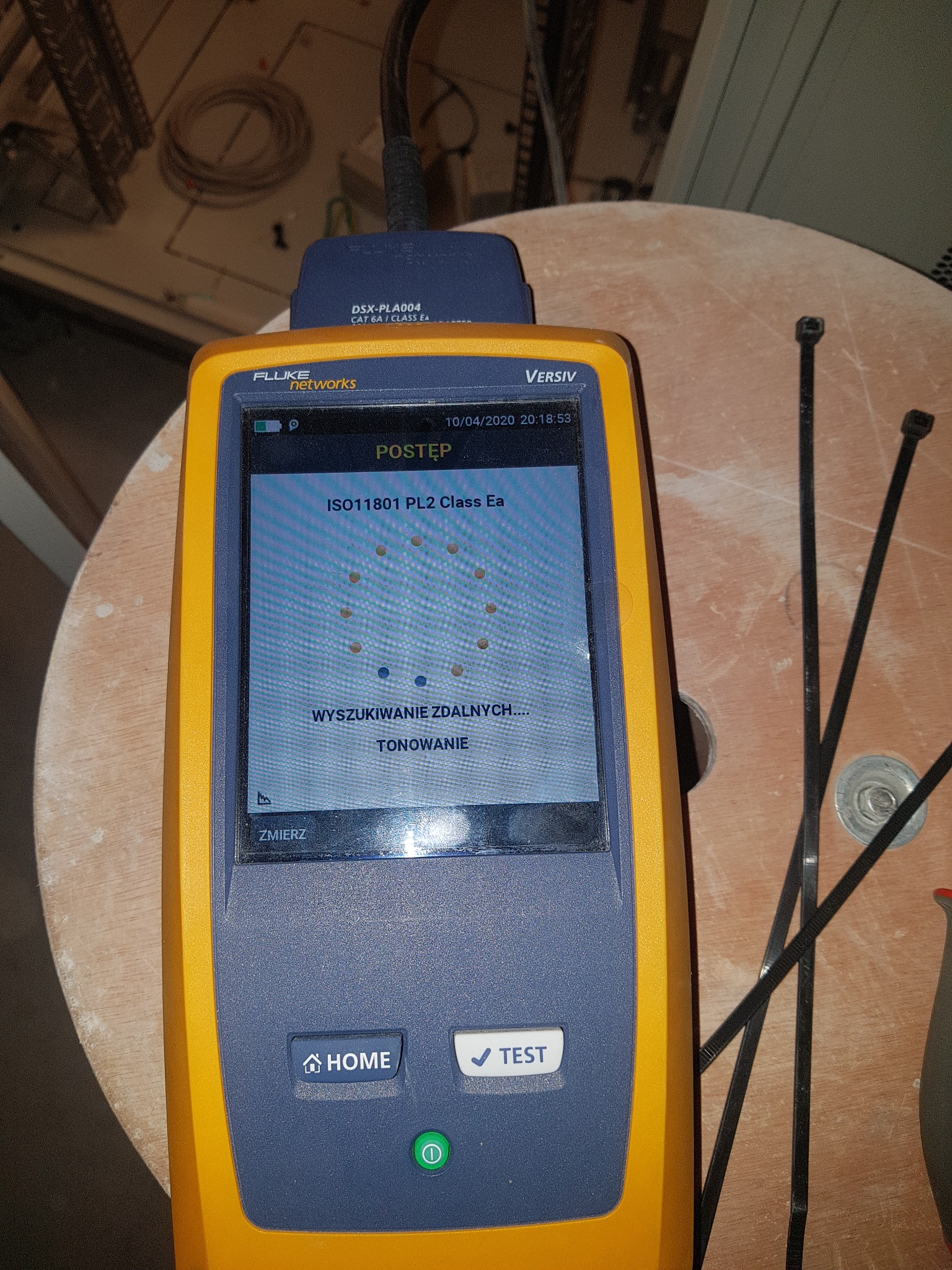 Miernik Fluke Networks Versiv wyświetlający wynik testu ISO11801 PL2 Class Ea podczas wyszukiwania zdalnych punktów, leżący na drewnianej powierzchni z opaskami zaciskowymi.