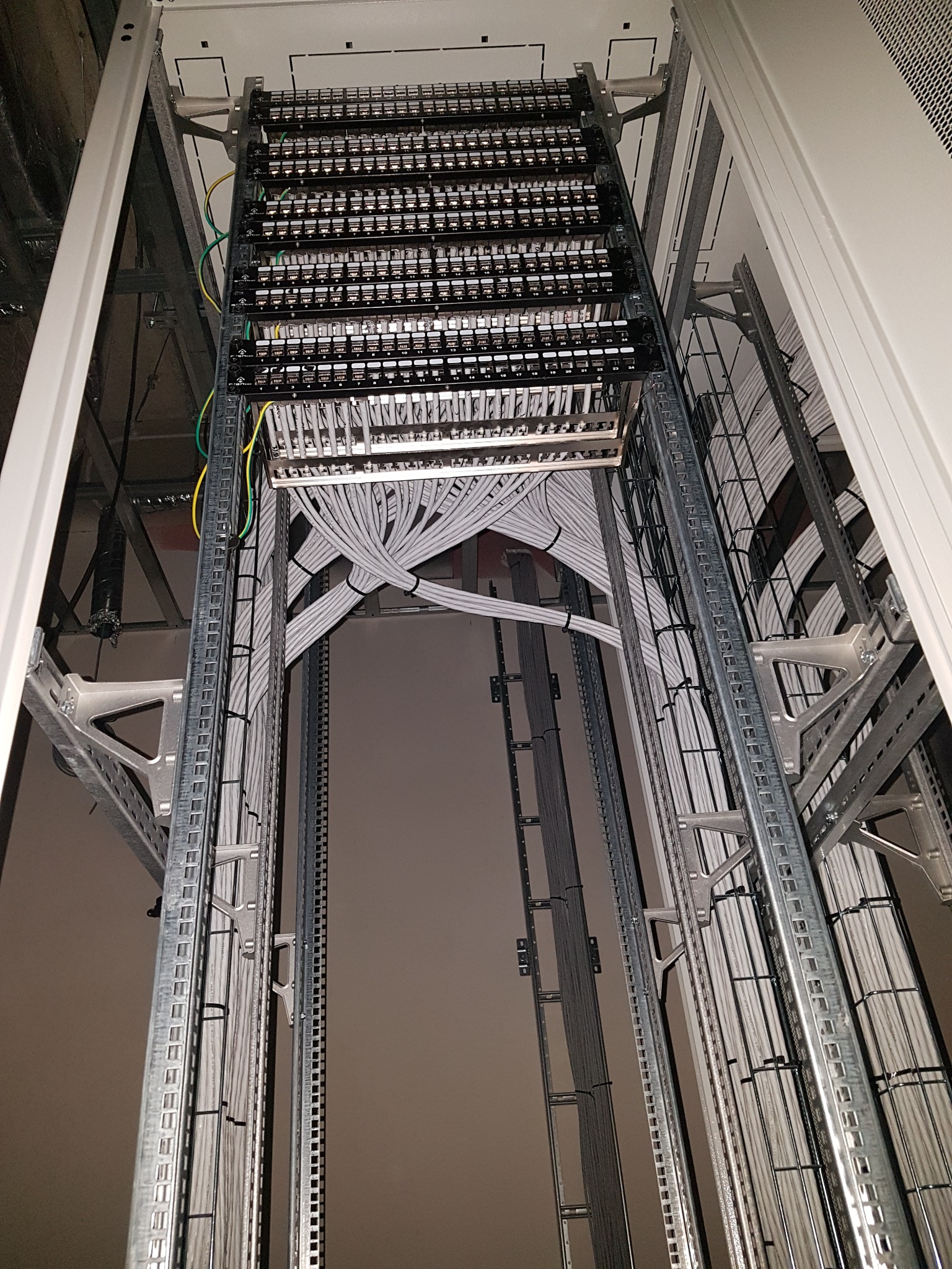 Widok z dołu na szafę krosowniczą z uporządkowanymi kablami sieciowymi, zamocowanymi na metalowych szynach i drabinkach kablowych w serwerowni.