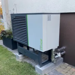 Zewnętrzna jednostka pompy ciepła Calla Verde zainstalowana przy budynku, z widocznymi izolowanymi rurami i donicą z roślinami obok.