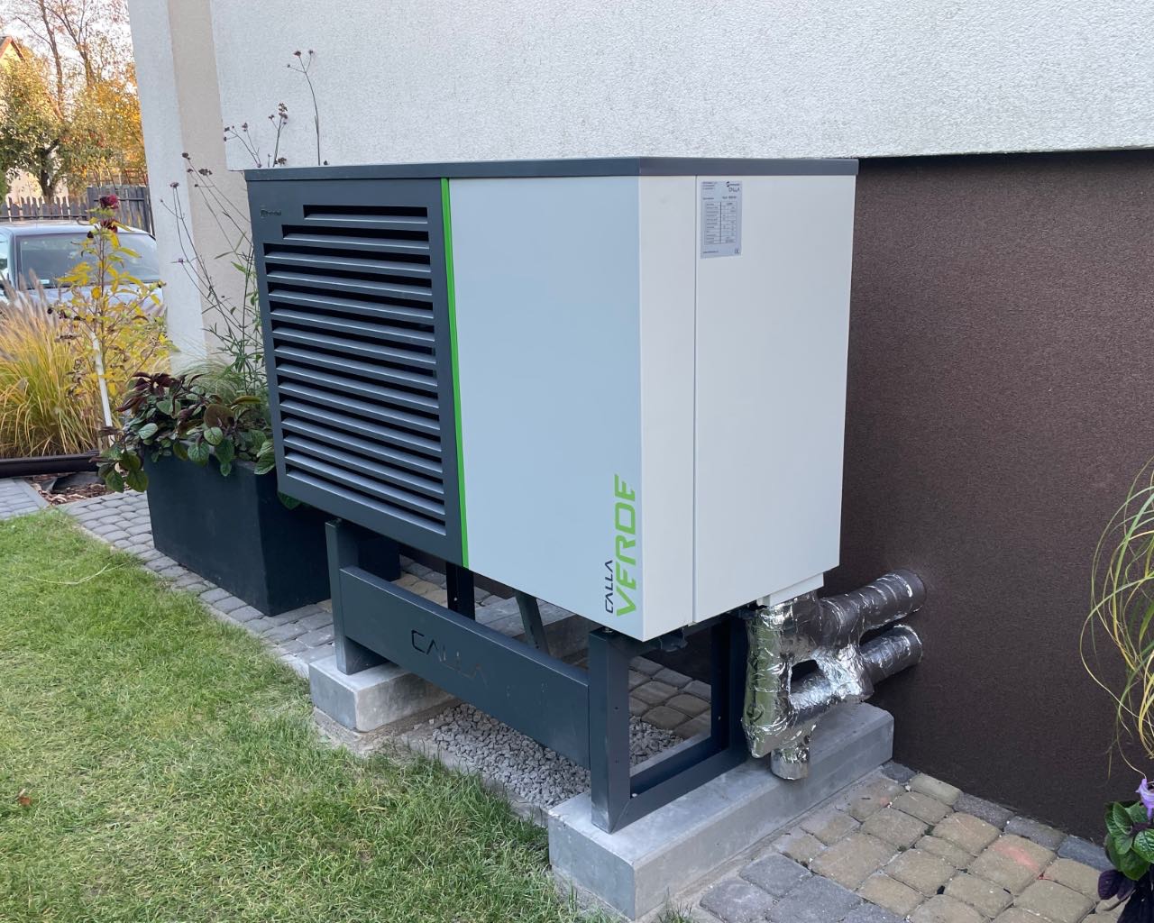 Zewnętrzna jednostka pompy ciepła Calla Verde zainstalowana przy budynku, z widocznymi izolowanymi rurami i donicą z roślinami obok.