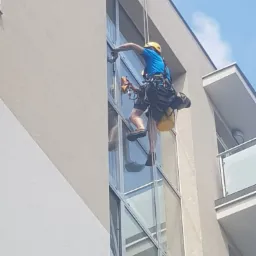 Pracownik w uprzęży alpinistycznej, z żółtym kaskiem, myje okna na wysokości w nowoczesnym budynku biurowym, używając specjalistycznego sprzętu do czyszczenia szyb.