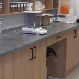 SYMBIOSYSTEM - Wnętrze laboratorium z blatem roboczym, wagą analityczną Ohaus, pojemnikami z substancjami i odkurzaczem przemysłowym w tle.