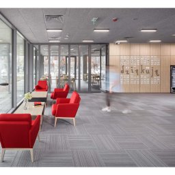 SYMBIOSYSTEM - Nowoczesne wnętrze biurowe z czerwonymi fotelami, szarą wykładziną dywanową w geometryczny wzór i ścianą z drewnianymi panelami z napisem 'dziękuję' i logotypami firm. Widoczne duże okna, lampa...