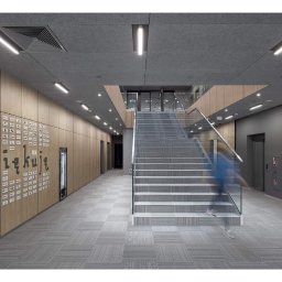 SYMBIOSYSTEM - Wnętrze nowoczesnego biura z jasną podłogą w kratę, drewnianymi panelami na ścianach z logo firm, schodami z szklaną balustradą i rozmytą sylwetką osoby schodzącej na dół.