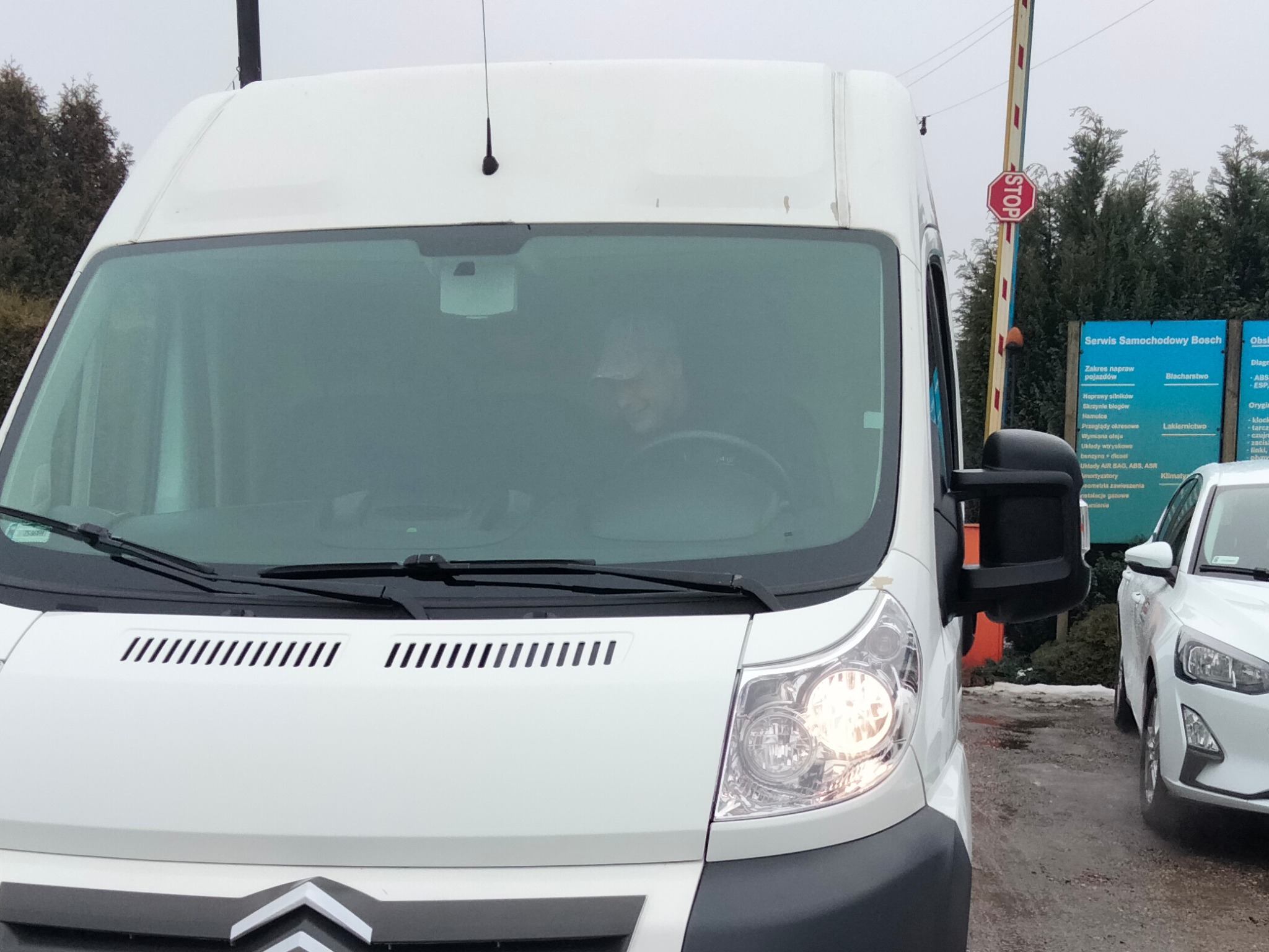 Biały samochód dostawczy marki Citroen, zaparkowany przed serwisem samochodowym Bosch, z widocznym kierowcą za kierownicą i znakiem STOP w tle.
