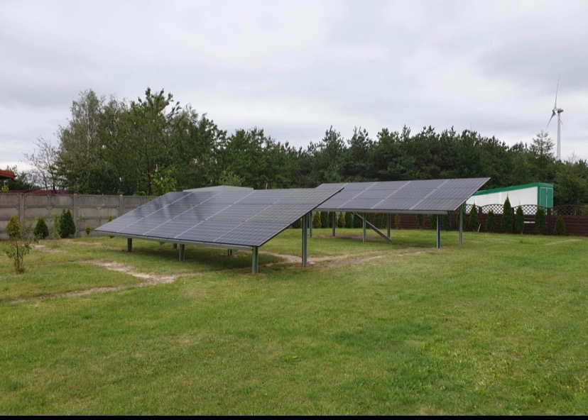 Dwa rzędy paneli fotowoltaicznych na metalowych konstrukcjach, ustawione na trawniku w ogrodzie, widoczny betonowy płot, drzewa i wiatrak w tle.