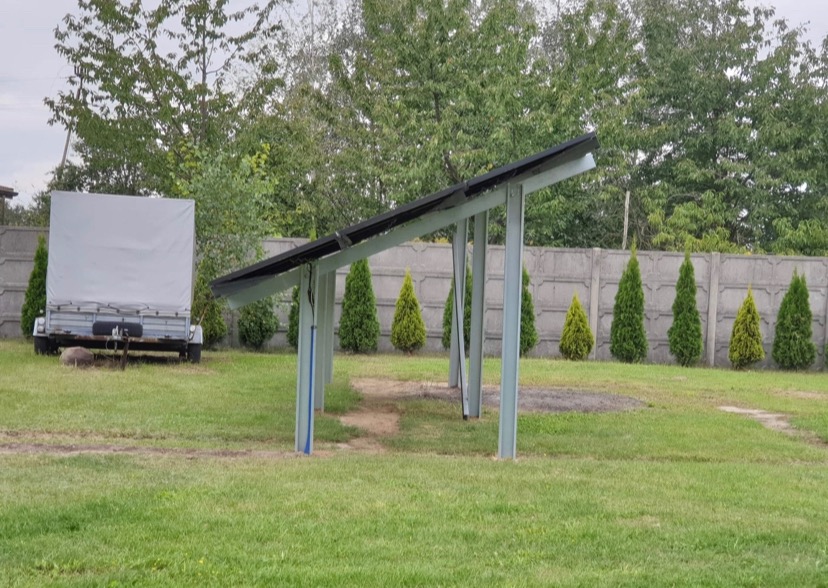 Zadaszenie panelami słonecznymi na metalowej konstrukcji w ogrodzie, widoczna przyczepka z plandeką i betonowe ogrodzenie w tle.
