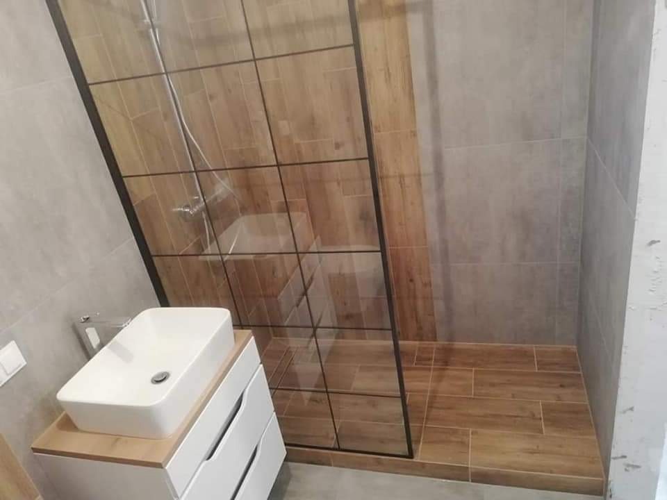 Nowoczesna łazienka z kabiną prysznicową typu walk-in z czarnymi profilami, umywalką nablatową na szafce z szufladami oraz płytkami imitującymi drewno na podłodze i ścianie w kabinie.