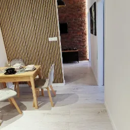 Wnętrze mieszkania z drewnianą ścianką działową z lameli, stołem z nakryciem, ścianą z czerwonej cegły i podłogą z płytek imitujących drewno.