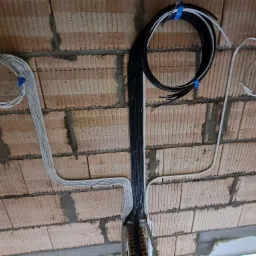 Wiązki przewodów elektrycznych (białe i czarne) przytwierdzone niebieską taśmą do surowego muru z cegły dziurawki w trakcie montażu.