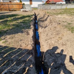 Elektro-Lasiu - Wykop w ziemi z ułożonymi niebieskimi rurami osłonowymi, widoczne domy w tle, cień osoby fotografującej w prawym dolnym rogu.